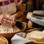 Manos muelen maíz nixtamalizado en un metate junto a tortillas recién hechas.
