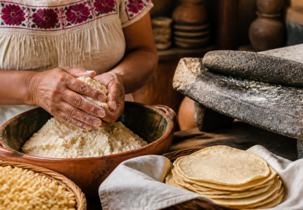 Manos muelen maíz nixtamalizado en un metate junto a tortillas recién hechas.