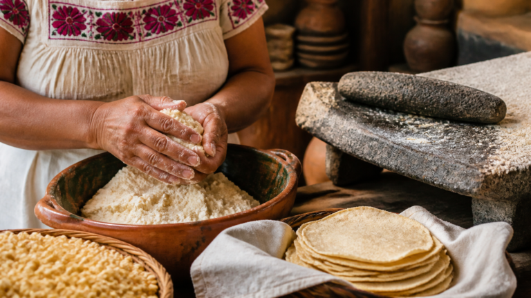 Manos muelen maíz nixtamalizado en un metate junto a tortillas recién hechas.